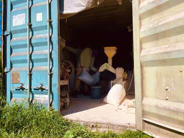 Eine Person arbeitet in einem Frachtcontainer, bedient Maschinen und hantiert mit einem großen weißen Sack. Säcke und Ausrüstung liegen verstreut herum; das Sonnenlicht beleuchtet die Szene teilweise.