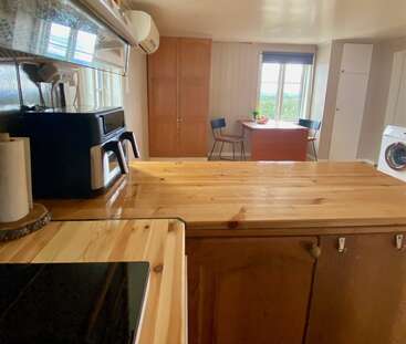 Esta imagen muestra una acogedora cocina con encimeras de madera, electrodomésticos modernos, una mesa de comedor con dos sillas, una ventana y una lavadora al fondo.