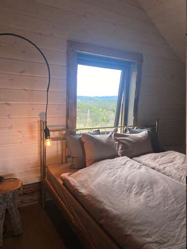 Acogedor dormitorio rústico con una cama de metal, ropa de cama en tonos neutros e iluminación tenue. Un gran ventanal ofrece vistas a un pintoresco paisaje boscoso. Las paredes de madera realzan el ambiente cálido.