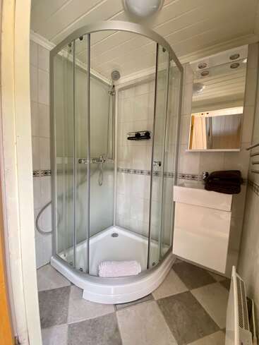 Se trata de un cuarto de baño pequeño y moderno que cuenta con una ducha en esquina con puertas de cristal, un lavabo blanco, un espejo, toallas marrones y un suelo de baldosas a cuadros.