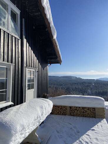 Una cabaña de madera con el tejado cubierto de nieve y un banco, con leña apilada debajo, domina un extenso paisaje forestal invernal bajo un cielo azul y despejado en un día soleado.