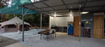 Esta imagen muestra una cocina y un comedor cubiertos al aire libre con mesa y sillas, una tienda cercana, un fregadero, puertas y rodeados de árboles.