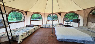 Dentro de una espaciosa tienda redonda con suelo de madera hay varias camas bien dispuestas, ropa de cama acogedora, almohadas, estanterías con provisiones y grandes ventanales que muestran la verde naturaleza del exterior.