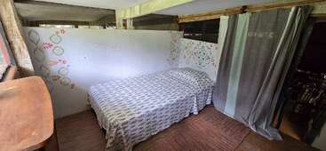 Un lit simple confortable avec une literie à motifs est placé à côté d'un mur décoré, d'un sol en bois, d'une table en bois et de rideaux séparant la pièce d'un autre espace.