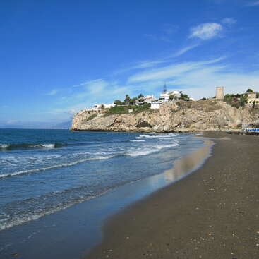 The image depicts a serene beach scene with a cliffside in the background, featuring a few buildings and a vibrant blue sky with wispy clouds.