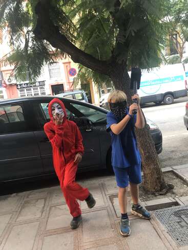 Two children wearing masks stand on a sidewalk, one in a red jumpsuit and the other in blue, with a car and tree behind them.