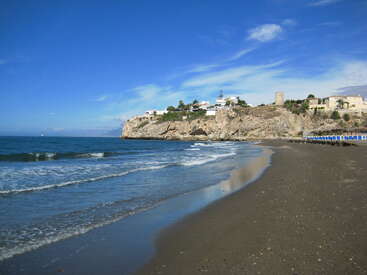 The image depicts a serene beach scene with a cliffside in the background, featuring a few buildings and a vibrant blue sky with wispy clouds.