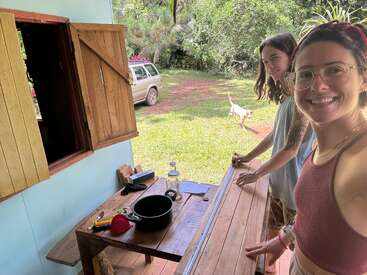 Deux femmes sourient en travaillant sur un projet en bois à l'extérieur d'une maison rustique. Des outils et un pot sont posés sur la table. Un chien et une voiture se trouvent à l'arrière-plan.