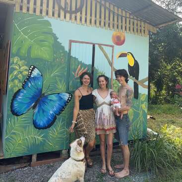 Três adultos, um bebê e um cachorro ficam felizes em frente a um mural pintado com cores vivas, com uma borboleta azul, plantas tropicais exuberantes e um tucano.