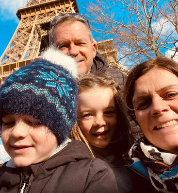 Uma família de quatro pessoas posa sorrindo em frente à Torre Eiffel num dia ensolarado. As crianças estão bem agasalhadas, com gorros e casacos quentinhos.
