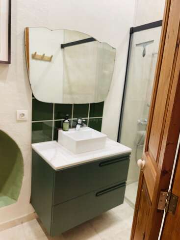 Esta imagem mostra um banheiro pequeno com um lavatório verde, pia branca, um espelho irregular bem original, box de vidro, porta de madeira e um revestimento de azulejos verdes. Moderno e arrumado.
