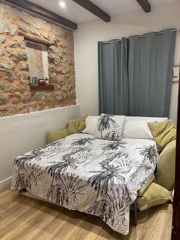 Um quarto aconchegante tem uma parede com detalhes em pedra, vigas de madeira, uma cama com roupa de cama com estampas tropicais, almofadas verdes, cortinas cinzas e um piso de madeira aconchegante, criando um charme rústico.