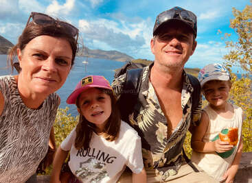 Uma família de quatro pessoas sorri durante uma aventura ao ar livre num dia ensolarado, com águas azuis e montanhas verdes ao fundo, registrando uma lembrança feliz das férias que passaram juntos.
