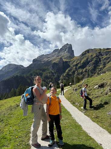 A group hikes along a winding mountain trail under a bright, partly cloudy sky, surrounded by green hills and dramatic rocky peaks, enjoying the outdoors.