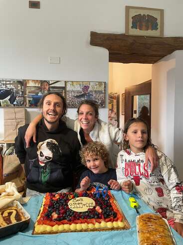 A happy family gathers around a fruit-topped birthday cake, smiling joyfully. The table has pastries, and colorful decorations enhance the celebration’s joyful, warm atmosphere at home.