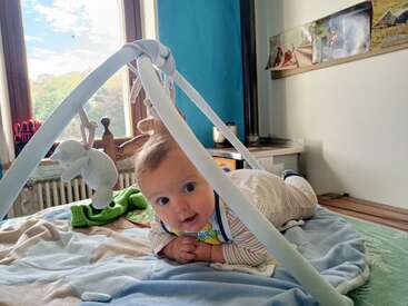 A baby lies on a bed under a play gym, smiling. Sunlight streams through large windows, creating a cozy, cheerful atmosphere in this comfortable room.