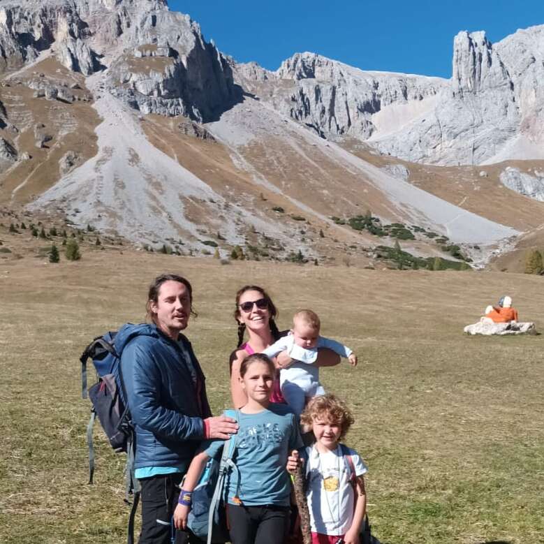 A family of five, dressed for hiking, poses happily in front of rocky mountains and blue sky, enjoying a beautiful sunny day in nature.