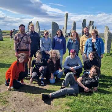 Un groupe de treize personnes pose joyeusement en plein air devant d'anciennes pierres debout, sous un ciel d'un bleu éclatant avec des nuages épars, profitant d'une journée ensoleillée.