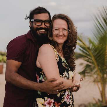 The image depicts a man and woman embracing on a beach, with the man wearing a maroon shirt and the woman in a black floral dress, set against a palm tree backdrop.