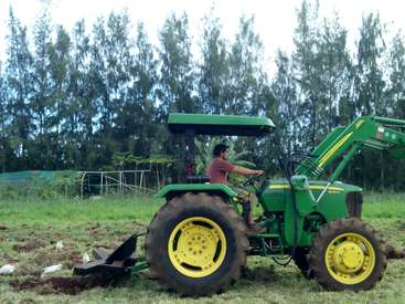 A man is operating a green John Deere tractor with a plow attachment in a field, surrounded by trees and a cloudy sky.