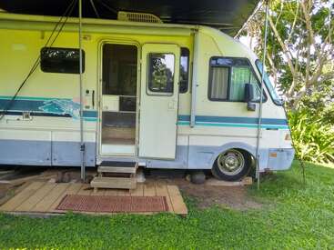 A parked RV with an open door sits on grass, shaded by trees. Wooden steps and a doormat lead up to the entrance, creating a cozy atmosphere.