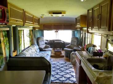 This image shows the interior of an RV, featuring a cozy seating area, kitchen sink, wooden cabinets, two front seats, curtains, a table, and decorative rug.