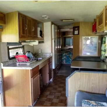 This image shows the interior of an RV featuring a compact kitchen, cabinets, countertop, sink, dining area with table, wood accents, and cozy seating.