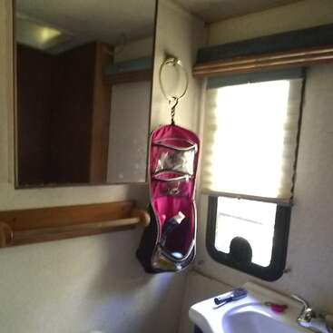This image shows a small bathroom with a mirror, a towel rack, a window with blinds, a sink, and a pink hanging organizer containing toiletries.