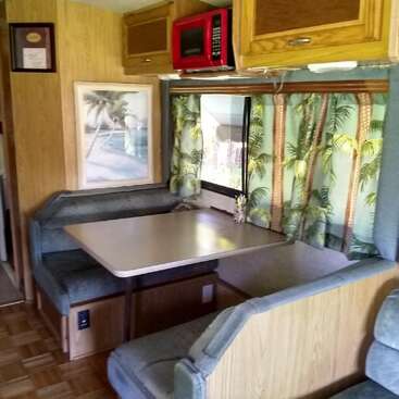 This image shows a cozy RV dining area with a table, cushioned bench seats, tropical palm tree curtains, a red microwave, wood cabinets, and a beach painting.