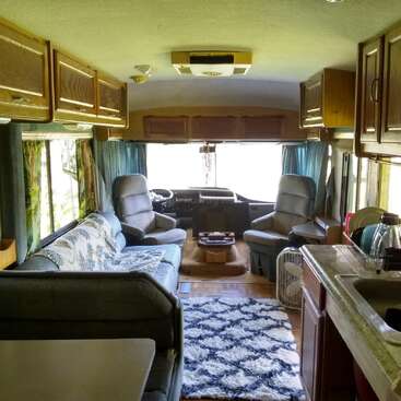 This image shows the cozy interior of an RV, complete with cushioned chairs, a sofa, kitchen, windows, storage cabinets, a patterned rug, and natural light.