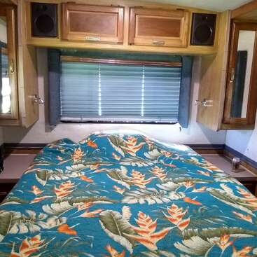 This image shows a cozy RV bedroom with wood cabinets, a large window with blinds, and a bed covered in a colorful tropical floral quilt.