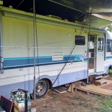 A motorhome is parked with an awning overhead, wooden steps lead to the door. A suitcase and outdoor mat are visible, suggesting a temporary outdoor setup.