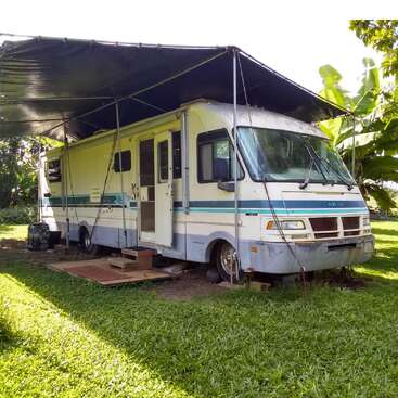 An old motorhome is parked on green grass, covered by a large black tarp canopy for shade. Steps, mats, and plants create a cozy outdoor setup.