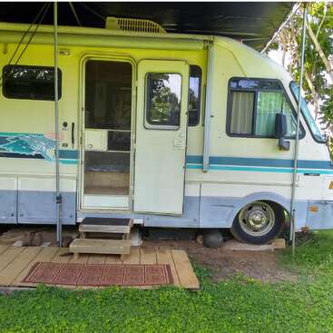 A vintage RV sits parked on grass, with wooden steps and a welcome mat leading to its open door. Outdoor canopy provides shade, surrounded by greenery.