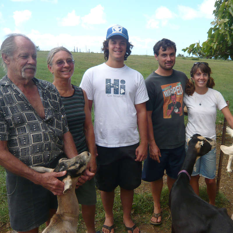 The image shows a group of five people standing in a grassy field, with two goats and one dog, all facing the camera and smiling.