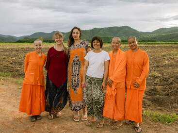 Seis mujeres sonríen juntas en el exterior; tres llevan túnicas budistas naranjas y las otras, vestidos de colores. Detrás de ellas hay campos y lejanas colinas verdes.