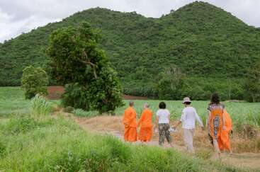 Un grupo de personas, entre ellas monjes con túnicas naranjas, caminan por un sendero rural rodeado de exuberante vegetación y colinas bajo un cielo nublado. Un entorno tranquilo y sereno.