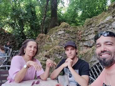 Três pessoas sentam-se em uma mesa ao ar livre cercada por paredes de pedra e vegetação. Uma pessoa sorri para a câmera, enquanto as outras parecem neutras ou pensativas.