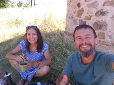 Um homem e uma mulher estão sorrindo, sentados na grama seca ao lado de um muro de pedra, tomando uma bebida juntos. Parece ensolarado e relaxado.