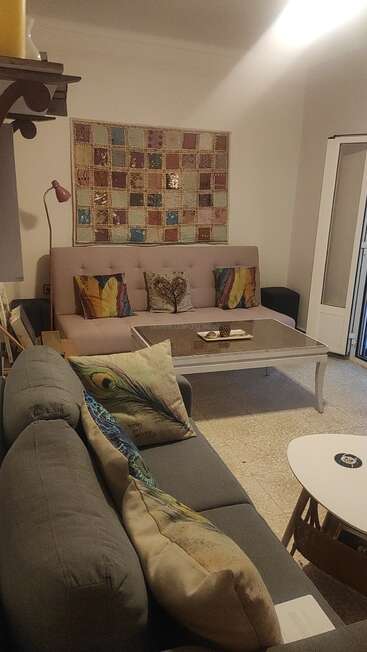 Uma sala de estar aconchegante com almofadas coloridas, dois sofás, uma decoração de parede em patchwork, mesas de centro, uma luminária de pé e uma iluminação ambiente quente, criando uma atmosfera acolhedora.