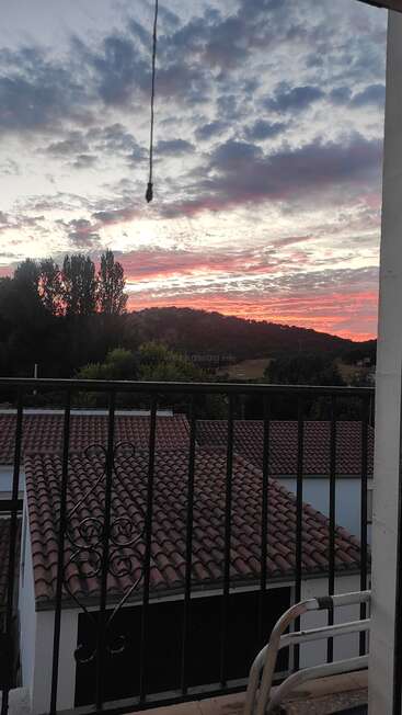 Um lindo céu ao pôr do sol pintado com nuvens rosa e roxas, visto de uma varanda, com vista para telhados, colinas, árvores verdes e um horizonte tranquilo.
