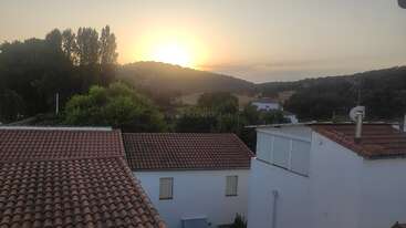 Um belo nascer do sol sobre colinas onduladas, com casas brancas com telhados vermelhos em primeiro plano, cercadas por árvores verdes exuberantes e uma cena rural tranquila.