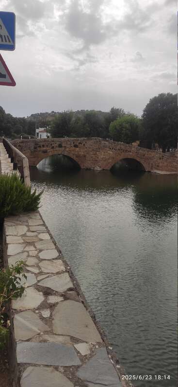 A rustic stone bridge with two arches crosses over a calm river, surrounded by greenery and trees, under a cloudy sky in a peaceful setting.