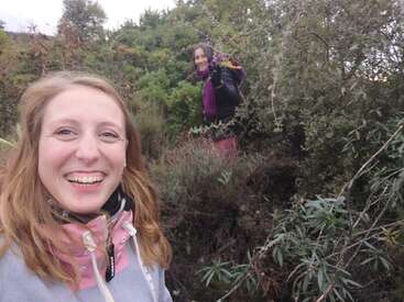 Deux femmes profitent d'une aventure en plein air dans une forêt luxuriante et verdoyante. L'une sourit à l'appareil photo tandis que l'autre se tient debout parmi les buissons, un bouquet de fleurs à la main.