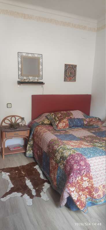 Ein gemütliches Schlafzimmer mit einer bunten Patchworkdecke, einem roten Kopfteil, einem kleinen Nachttisch aus Holz, einem Deko-Spiegel, Wandbildern und einem braun-weißen Fellteppich.