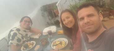 Drei Leute sitzen an einem Tisch im Freien, lächeln und genießen gemeinsam das Essen. Die Atmosphäre ist ungezwungen, um sie herum stehen Essen, Getränke und Pflanzen.