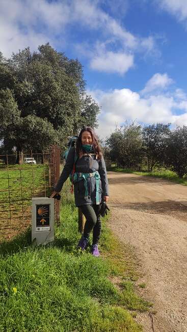 Eine Frau in Wanderkleidung und mit Rucksack steht an einem sonnigen Tag lächelnd neben einem Wegweiser des Jakobswegs, umgeben von einem Feldweg und viel Grün.