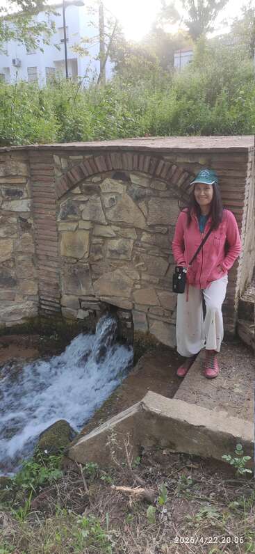 Eine Frau in einer rosa Jacke und einer grünen Mütze steht neben einer Steinmauer mit einem Torbogen, in der Nähe von fließendem Wasser, umgeben von Grün und Abendlicht.