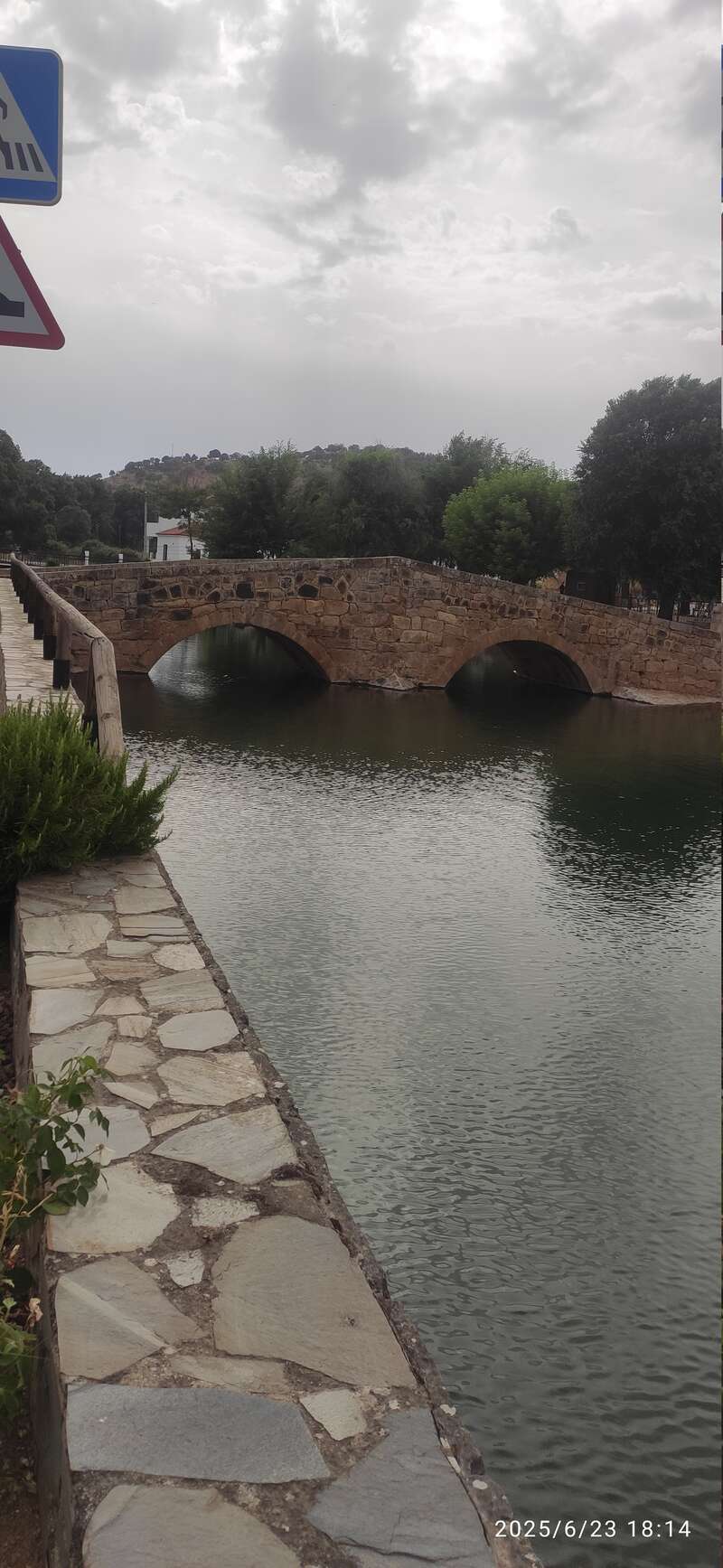 Uma ponte de pedra rústica com dois arcos atravessa um rio calmo, cercado por vegetação e árvores, sob um céu nublado em um ambiente tranquilo.
