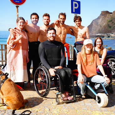 Um grupo de amigos, alguns sem camisa, outros sentados em cadeiras de rodas, sorrindo juntos perto de uma praia com montanhas, placas de estacionamento e um cachorro em primeiro plano.